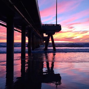 May include: A pier extends into the ocean at sunset. The sky is a blend of pink, purple, and orange hues. The pier's supports and structure are silhouetted against the colorful sky, with reflections in the wet sand.