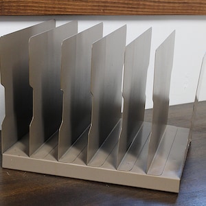 May include: A metal file organizer with six vertical dividers. The organizer is a muted silver color and sits on a rectangular base. The dividers have a notched design on the top edge.