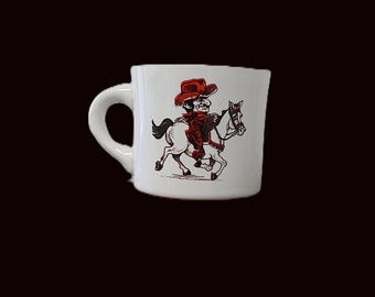 May include: White ceramic mug featuring a cartoon cowboy riding a horse. The cowboy wears a red hat and jacket, with the horse and rider in black and red ink. The mug has a curved handle.