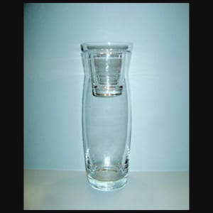 May include: A clear glass vase with a unique design, featuring a smaller, nested glass insert. The vase has a slender, elongated shape with a wide base and a slightly flared top. The glass is transparent, allowing light to pass through.