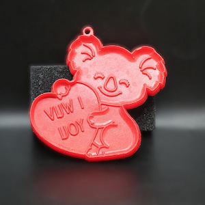 May include: Red koala-shaped ornament holding a heart with the text "VUW I UOY". The koala has a smiling face and a small loop at the top for hanging. The ornament is made of a solid, smooth material.