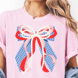 May include: A light pink t-shirt featuring a large bow design. The bow has a blue and white gingham base, red and pink ribbons, and strings of white pearls. The shirt is a casual style with rolled-up sleeves.