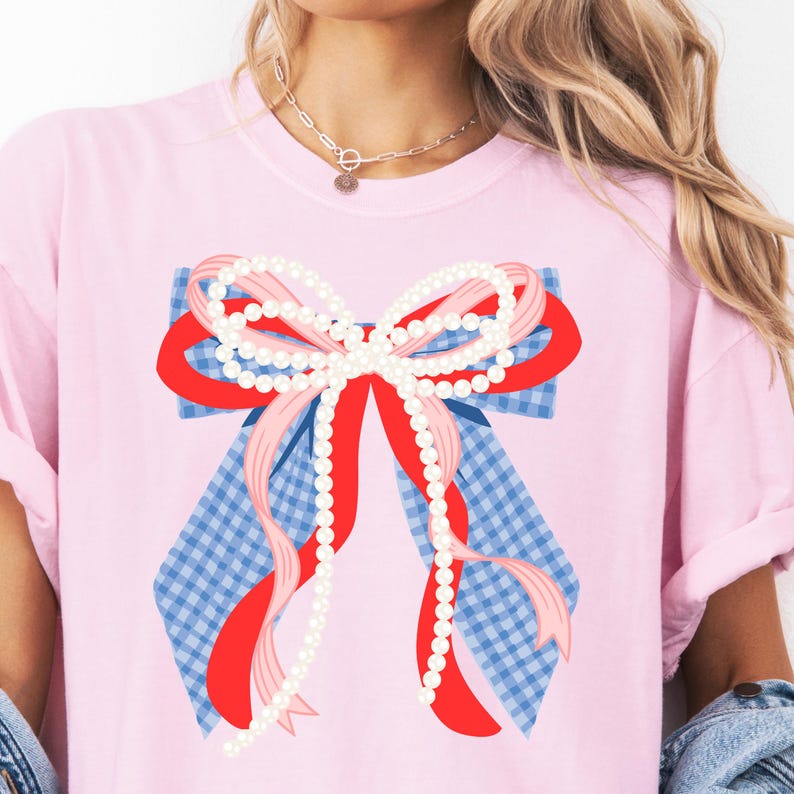 May include: A light pink t-shirt featuring a large bow design. The bow has a blue and white gingham base, red and pink ribbons, and strings of white pearls. The shirt is a casual style with rolled-up sleeves.