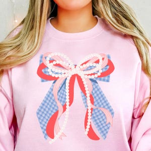 May include: A light pink sweatshirt featuring a large bow design. The bow has a blue and white gingham base, red and pink ribbons, and is adorned with strings of white pearls. The sweatshirt is a solid light pink color.