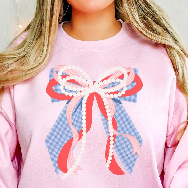 May include: A light pink sweatshirt featuring a large bow design. The bow has a blue and white gingham base, red and pink ribbons, and is adorned with strings of white pearls. The sweatshirt is a solid light pink color.