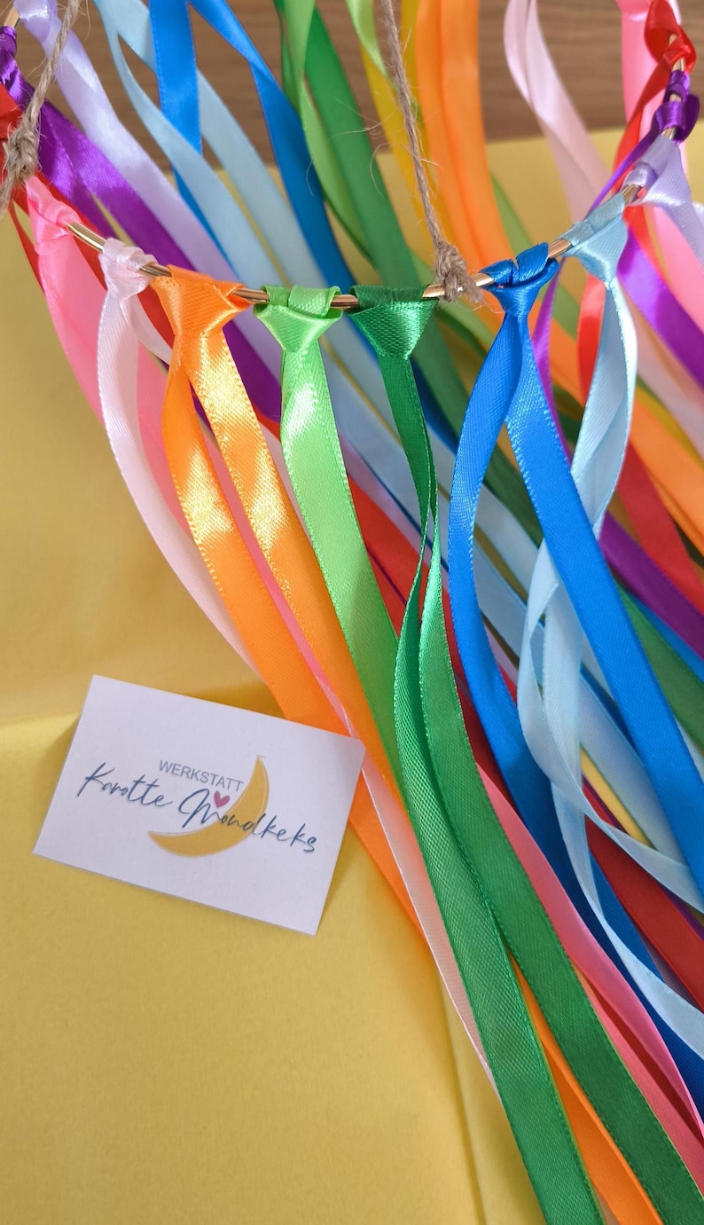 May include: A colorful ribbon wand with a gold hoop and a jute string for hanging. The ribbons are in various colors, including pink, orange, green, blue, and purple. A small card with text is visible.