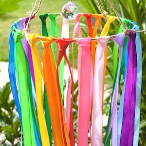 May include: A colorful wind chime with a rainbow of ribbons hanging from a circular frame. The ribbons are in shades of red, orange, yellow, green, blue, and purple. A crystal prism and wooden beads hang from the top.