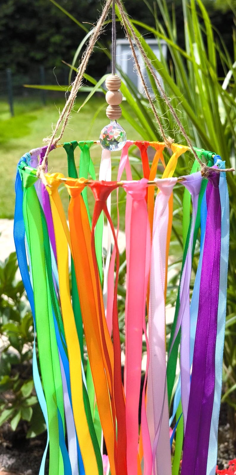 May include: A colorful wind chime with a rainbow of ribbons hanging from a circular frame. The ribbons are in shades of red, orange, yellow, green, blue, and purple. A crystal prism and wooden beads hang from the top.