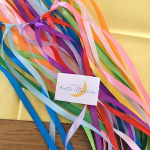 May include: A collection of colorful ribbons in various shades, including blue, green, yellow, orange, and purple. The ribbons are tied together at the top with a natural twine. A small card with text and a crescent moon graphic is attached.