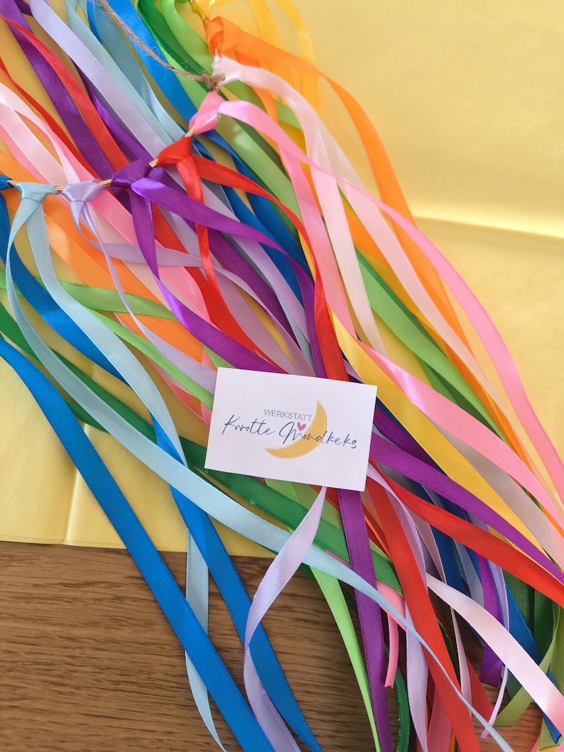 May include: A collection of colorful ribbons in various shades, including blue, green, yellow, orange, and purple. The ribbons are tied together at the top with a natural twine. A small card with text and a crescent moon graphic is attached.