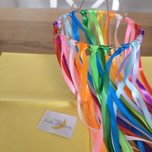 May include: A colorful mobile featuring a gold ring adorned with vibrant ribbons in shades of pink, orange, green, blue, and more. The ribbons hang down, creating a decorative effect. The mobile is suspended by twine.