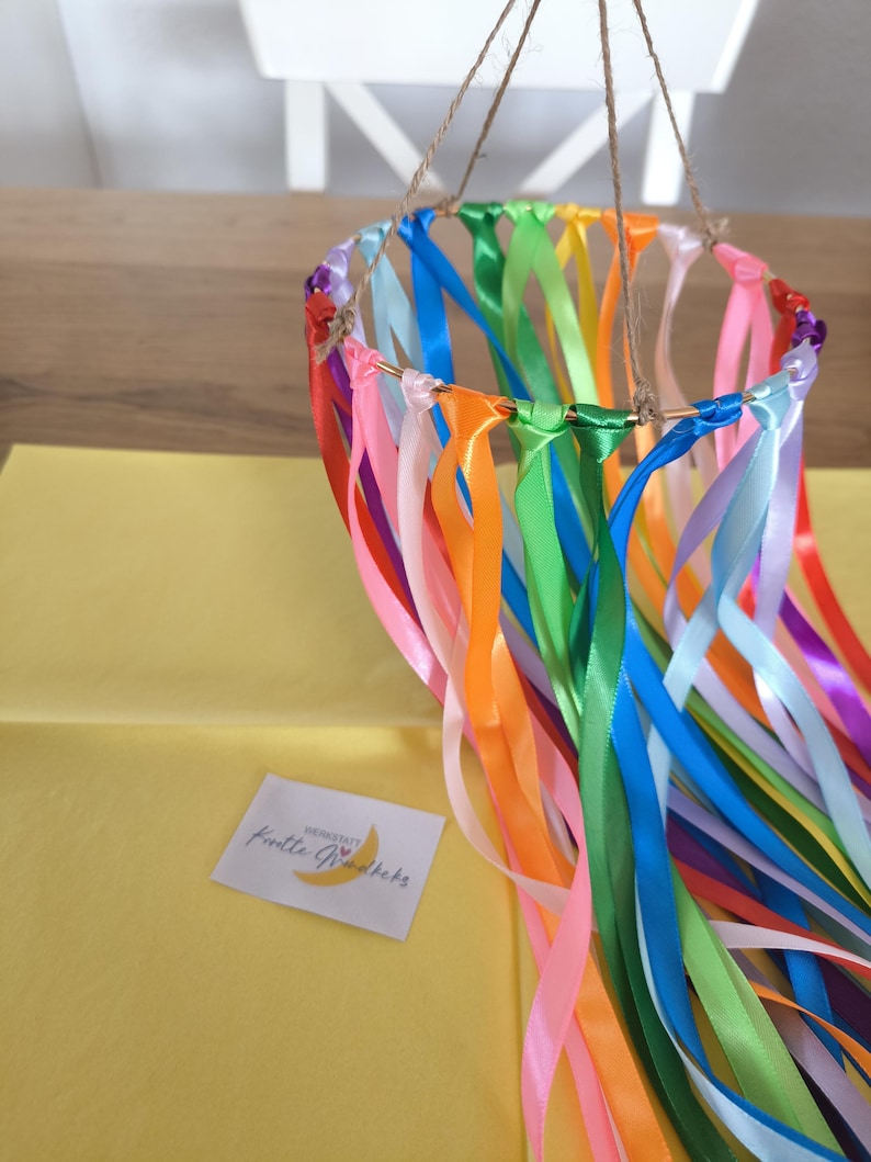 May include: A colorful mobile featuring a gold ring adorned with vibrant ribbons in shades of pink, orange, green, blue, and more. The ribbons hang down, creating a decorative effect. The mobile is suspended by twine.