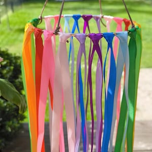 May include: A colorful wind chime with a rainbow of ribbons hanging from a circular frame. The ribbons are in shades of orange, pink, purple, blue, and green. The chime is suspended by a brown cord, creating a vibrant outdoor decoration.