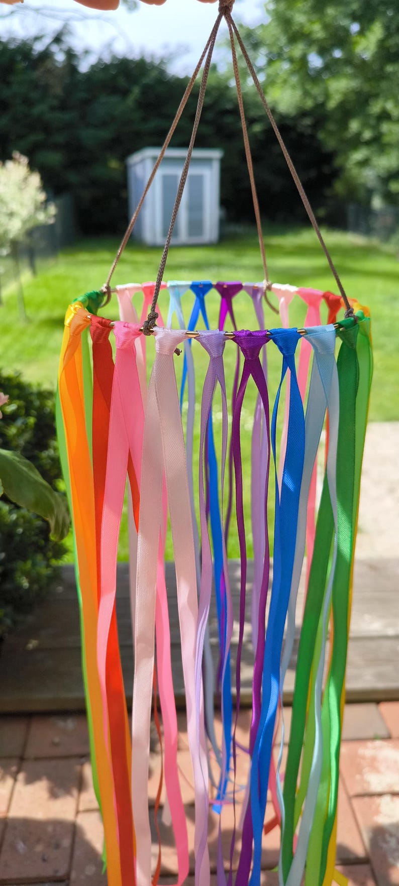 May include: A colorful wind chime with a rainbow of ribbons hanging from a circular frame. The ribbons are in shades of orange, pink, purple, blue, and green. The chime is suspended by a brown cord, creating a vibrant outdoor decoration.