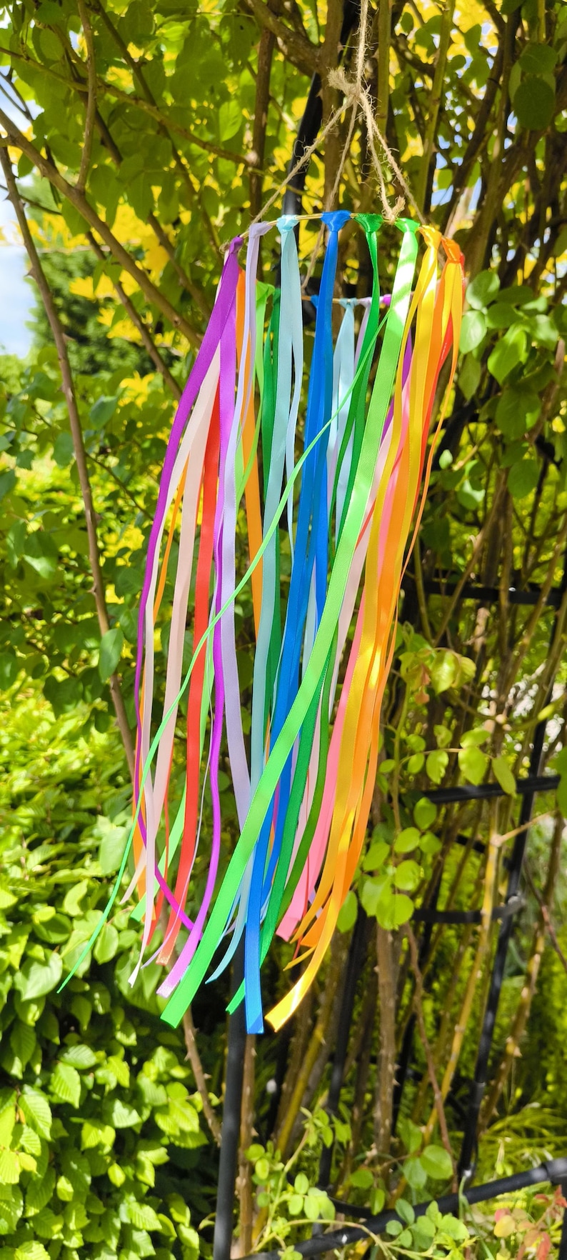 May include: A colorful wind chime with long, flowing ribbons in shades of pink, orange, yellow, green, blue, and purple. The ribbons hang from a circular frame suspended by a natural twine, creating a vibrant outdoor decoration.