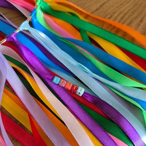 May include: A colorful ribbon wand with a gold ring and the name "Lottie" spelled out in letter beads. The ribbons are in various colors, including pink, orange, yellow, green, blue, purple, and white.