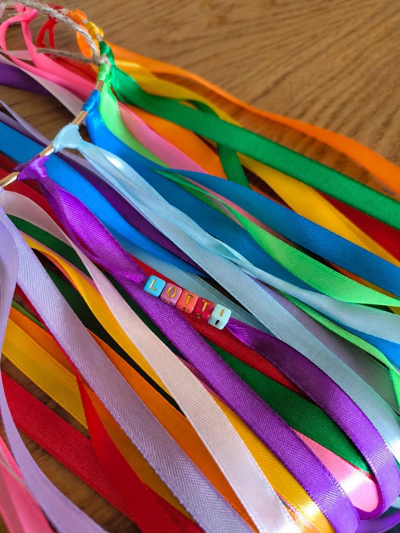 May include: A colorful ribbon wand with a gold ring and the name "Lottie" spelled out in letter beads. The ribbons are in various colors, including pink, orange, yellow, green, blue, purple, and white.