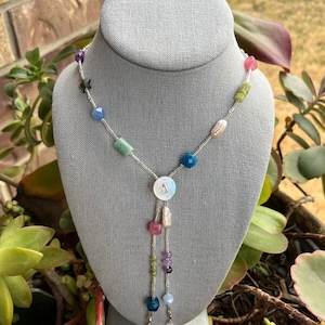 May include: A colorful beaded necklace with a silver chain and a button closure. The necklace features a variety of gemstones in shades of blue, green, pink, and purple. The necklace is displayed on a gray mannequin.
