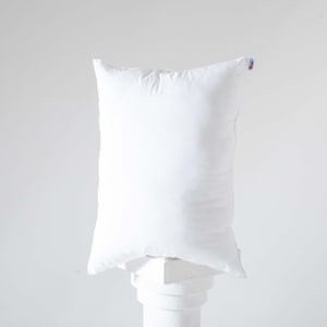 May include: A white pillow with a white tag on the top right corner. The pillow is sitting on a white pedestal.