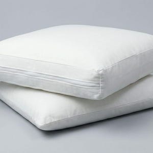 May include: Two square, white pillows stacked on top of each other. The top pillow has a zipper along one edge. The pillows are on a light gray background. These pillows are suitable for home decor.