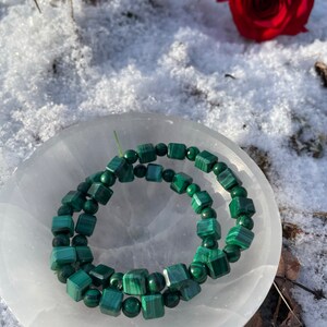 May include: A green malachite beaded bracelet on a white selenite charging plate. The beads are square shaped and have a natural, earthy look.