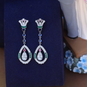 Peut inclure: Une paire de boucles d'oreilles pendantes en argent avec un motif en forme de larme. Les boucles d'oreilles présentent une couronne en haut, avec des pierres précieuses vertes, bleues et roses. La larme est entourée de pierres précieuses plus petites.