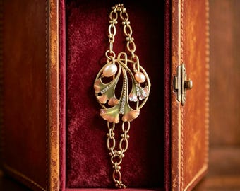 May include: A gold-toned bracelet featuring a central ginkgo leaf motif with green and pink enamel, small clear crystals, and two oval pearls. The bracelet is composed of a delicate chain with oval links and is displayed inside a brown box.