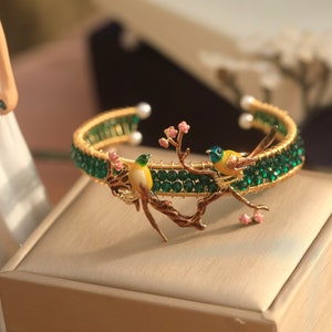 May include: A gold bracelet with green and pink accents features two yellow birds perched on a branch. The bracelet is made of wire and beads.