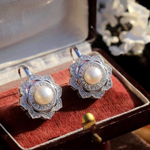 Peut inclure: Boucles d'oreilles en argent avec une perle centrale entourée de petites pierres précieuses transparentes. Les boucles d'oreilles ont une forme de fleur et sont présentées dans un écrin doublé de velours.