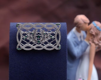 May include: A silver and green jeweled brooch with a decorative design. The brooch is on a blue velvet background.