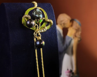 May include: A gold hair comb with a green and white floral design and three black pearls. The comb is on a blue velvet background.