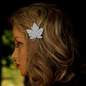 Op de afbeelding: Een zilverkleurige haarclip in de vorm van een esdoornblad, bezet met talrijke kleine, sprankelende kristallen. De clip staat tegen een achtergrond van golvend, lichtbruin haar.