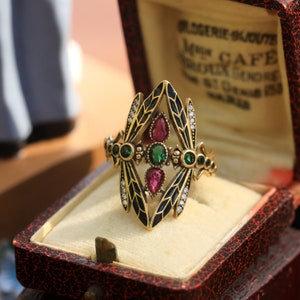 Puede incluir: Un anillo dorado con un diseño central con una gema verde flanqueada por dos gemas rojas. El anillo tiene detalles de esmalte negro y está adornado con pequeñas gemas blancas. Descansa en un joyero rojo y crema.