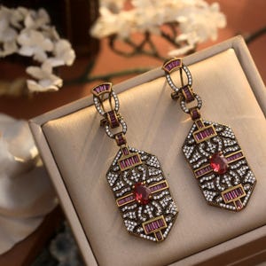 Peut inclure: Une paire de boucles d'oreilles dorées au design géométrique. Les boucles d'oreilles présentent un grand pendentif de forme octogonale avec une pierre précieuse rouge centrale entourée de petites pierres précieuses claires et roses. Les boucles d'oreilles sont sur un fond de velours blanc.