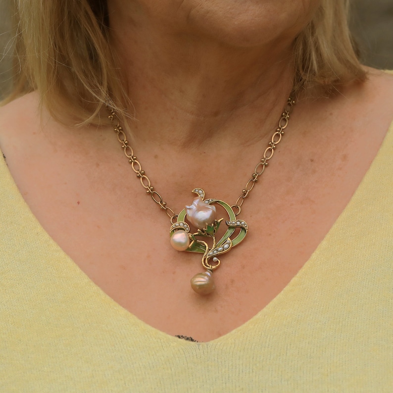 May include: A gold chain necklace with a pendant featuring a pink pearl flower with green enamel leaves and smaller pearls. The pendant hangs from a single larger pearl.