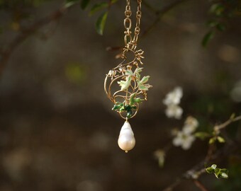 Può includere: Una collana color oro con un pendente con un design floreale con foglie di smalto verde e bianco e una perla a forma di goccia. La collana è sospesa da una delicata catena.