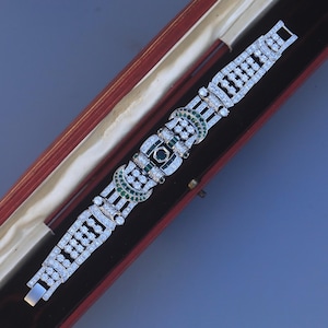 Może przedstawiać: Bransoleta w stylu Art Deco z oprawą z metalu w kolorze srebrnym, inkrustowana przezroczystymi i szmaragdowozielonymi kamieniami szlachetnymi. Bransoleta jest prezentowana w czerwonym pudełku na biżuterię z kremowym wnętrzem.