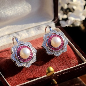 Può includere: Orecchini in argento con un design floreale. Ogni orecchino presenta una perla centrale, un anello di pietre rosa e un bordo di cristalli trasparenti. Gli orecchini sono esposti in un portagioie di velluto rosso.