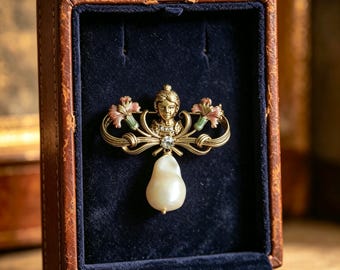 May include: A gold-toned brooch featuring a central human bust figure flanked by two pink and green enameled flowers. The piece includes a clear rhinestone accent and a large, irregular white baroque pearl drop, all displayed in a brown box.