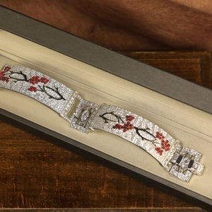 Puede incluir: Un brazalete de plata con un diseño floral engastado con piedras preciosas transparentes y rojas. El brazalete está en una caja de terciopelo negro.
