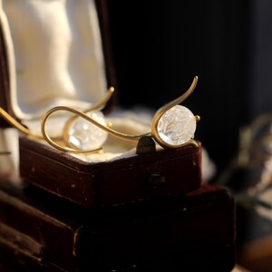 Peut inclure: Boucles d'oreilles dorées avec une sphère en cristal clair et facetté. Les boucles d'oreilles sont présentées dans un écrin à bijoux marron vintage ouvert avec un intérieur crème. Design incurvé.