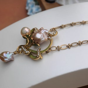 May include: A gold necklace with a pendant featuring a pink baroque pearl surrounded by smaller pearls and green enamel. The pendant is shaped like a flower with a delicate design. The necklace is made of a gold chain.