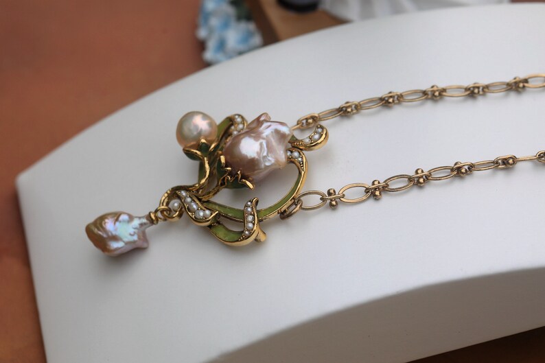 May include: A gold necklace with a pendant featuring a pink baroque pearl surrounded by smaller pearls and green enamel. The pendant is shaped like a flower with a delicate design. The necklace is made of a gold chain.