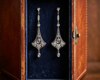 May include: A pair of silver-toned drop earrings featuring intricate filigree metalwork, clear pavé-set stones, and a central oval-cut stone, with a small teardrop-shaped stone dangling from the base, presented inside a brown leather box.