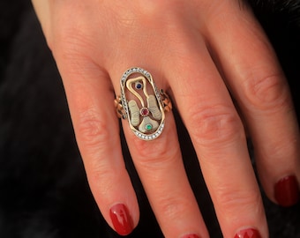 Op de afbeelding: Een gouden ring met een gedetailleerd ontwerp met een centraal motief van een gestileerde vogel met rode, blauwe en groene edelstenen. De ring is bezet met diamanten rondom de omtrek.
