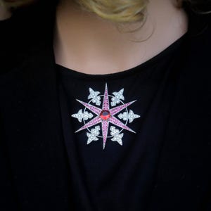 Op de afbeelding: Een stervormige broche met een centrale rode edelsteen en roze en witte accenten. De broche staat tegen een zwarte achtergrond, wat een opvallend contrast creëert. Een sieraad.