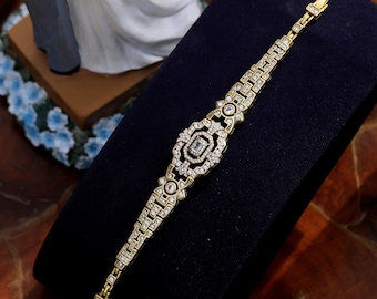May include: A gold bracelet with a large, square-shaped, clear crystal centerpiece surrounded by smaller crystals. The bracelet is made of a delicate chain with a geometric pattern.