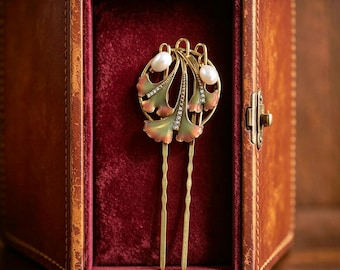 May include: A gold-toned hair fork featuring a circular frame with three ginkgo leaf-shaped elements. The leaves are finished with a gradient of green and pink enamel, accented by small clear crystals and two white oval pearls at the top.