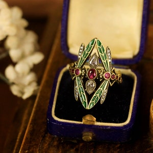 Op de afbeelding: Een gouden ring met een libellenontwerp. De vleugels van de libel zijn van groen emaille en het lichaam is bezet met rode en witte edelstenen.