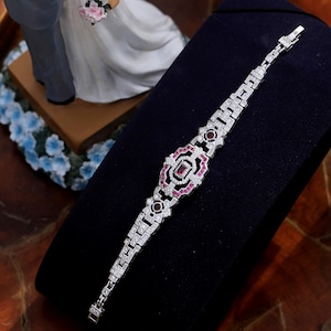 Peut inclure: Un bracelet en argent avec un motif géométrique mettant en valeur un carré central avec une pierre rose entourée de pierres roses plus petites. Le bracelet est serti de pierres claires.
